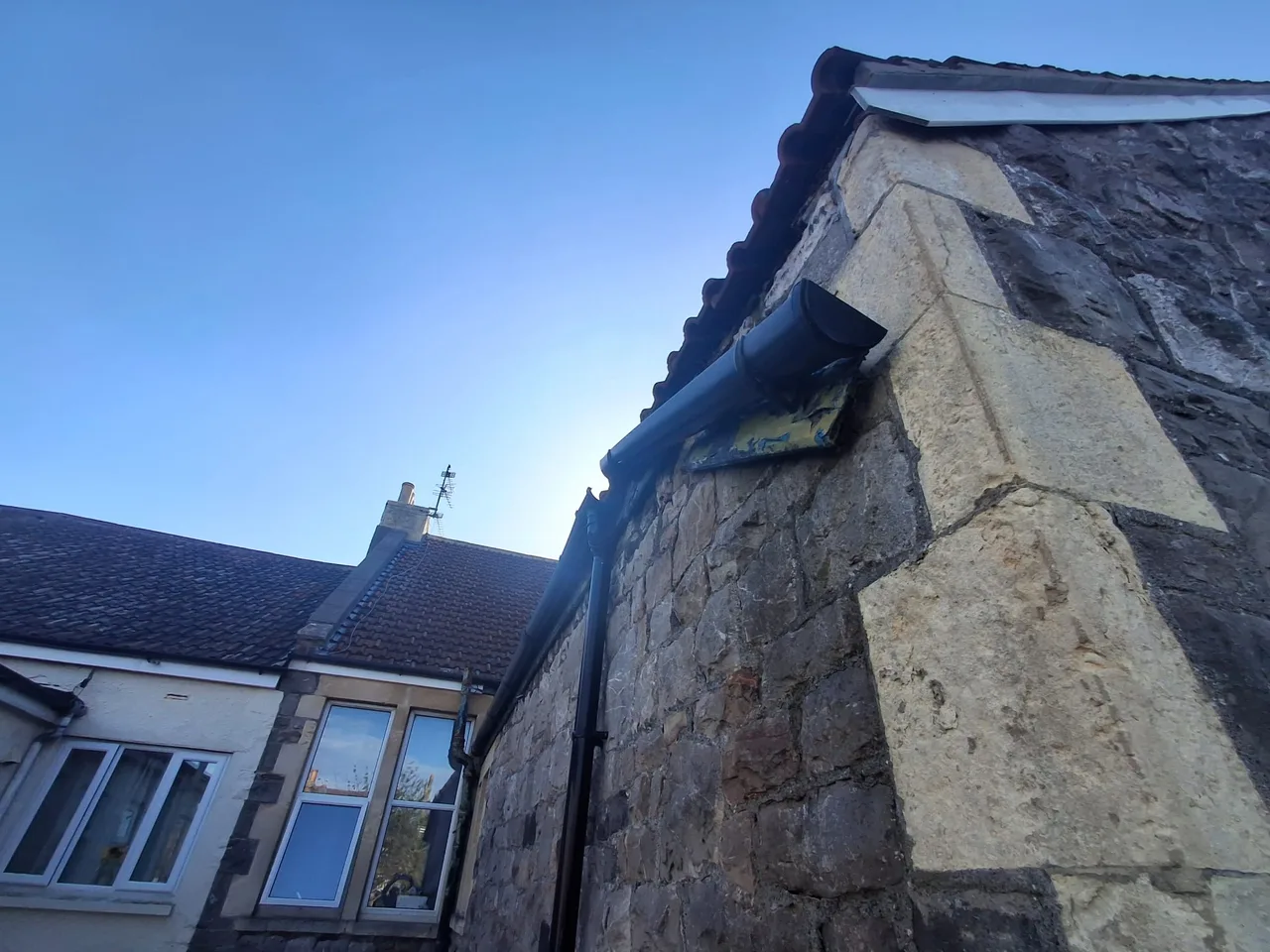 Peeling paint on the fascia behind a black gutter end above the corner of a stone house.