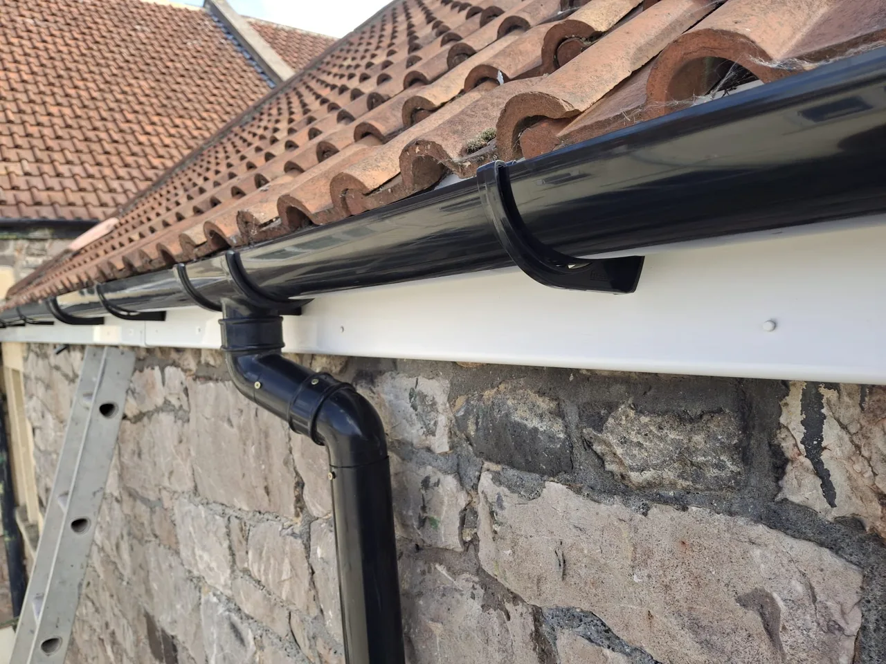 Black guttering and downpipe with white uPVC fascia installed by Lewis on a stone house